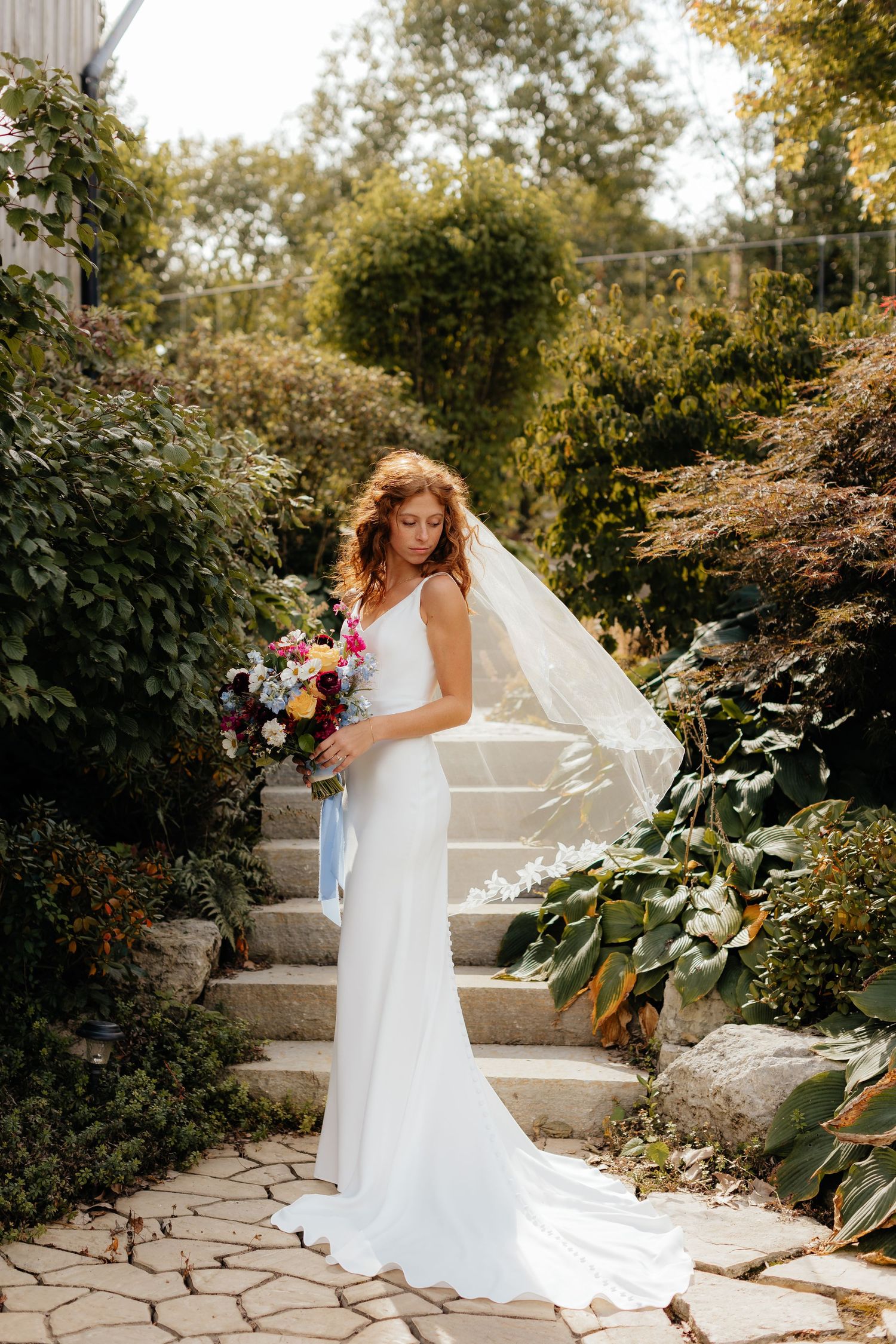 Bride in elegant white dress poses on garden steps with flowing veil and colorful bouquet.