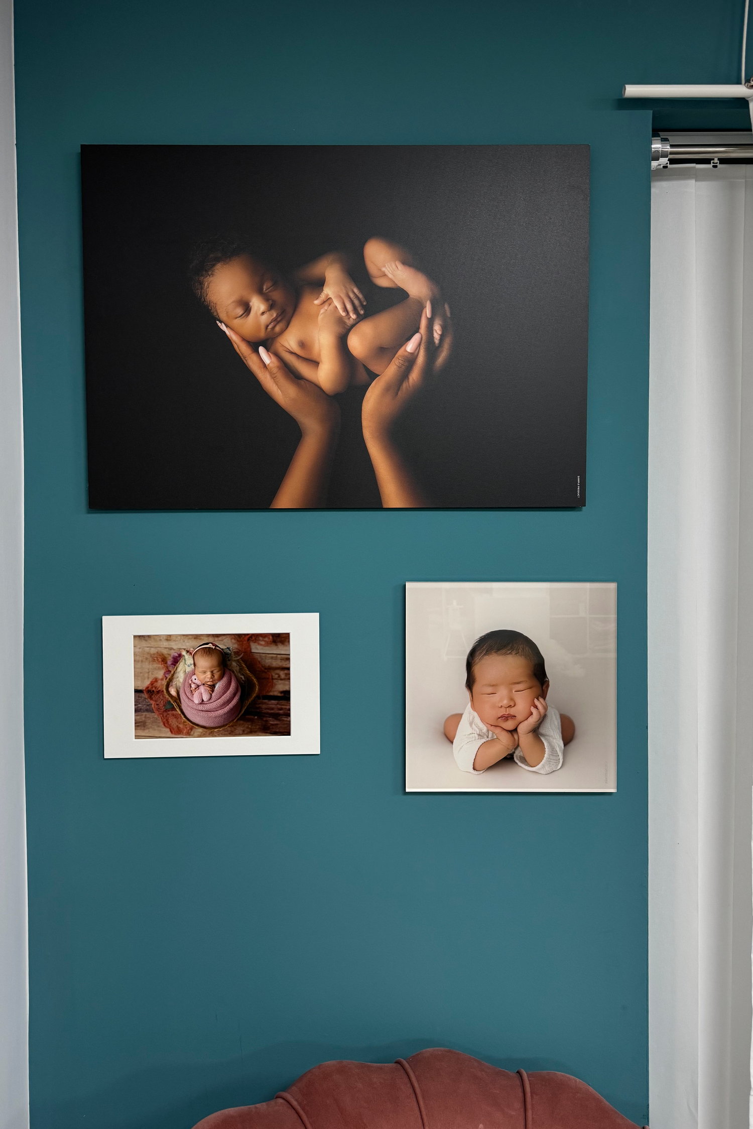Three portrait photographs arranged on a teal wall above a sofa, displaying artistic newborn photography in white frames.