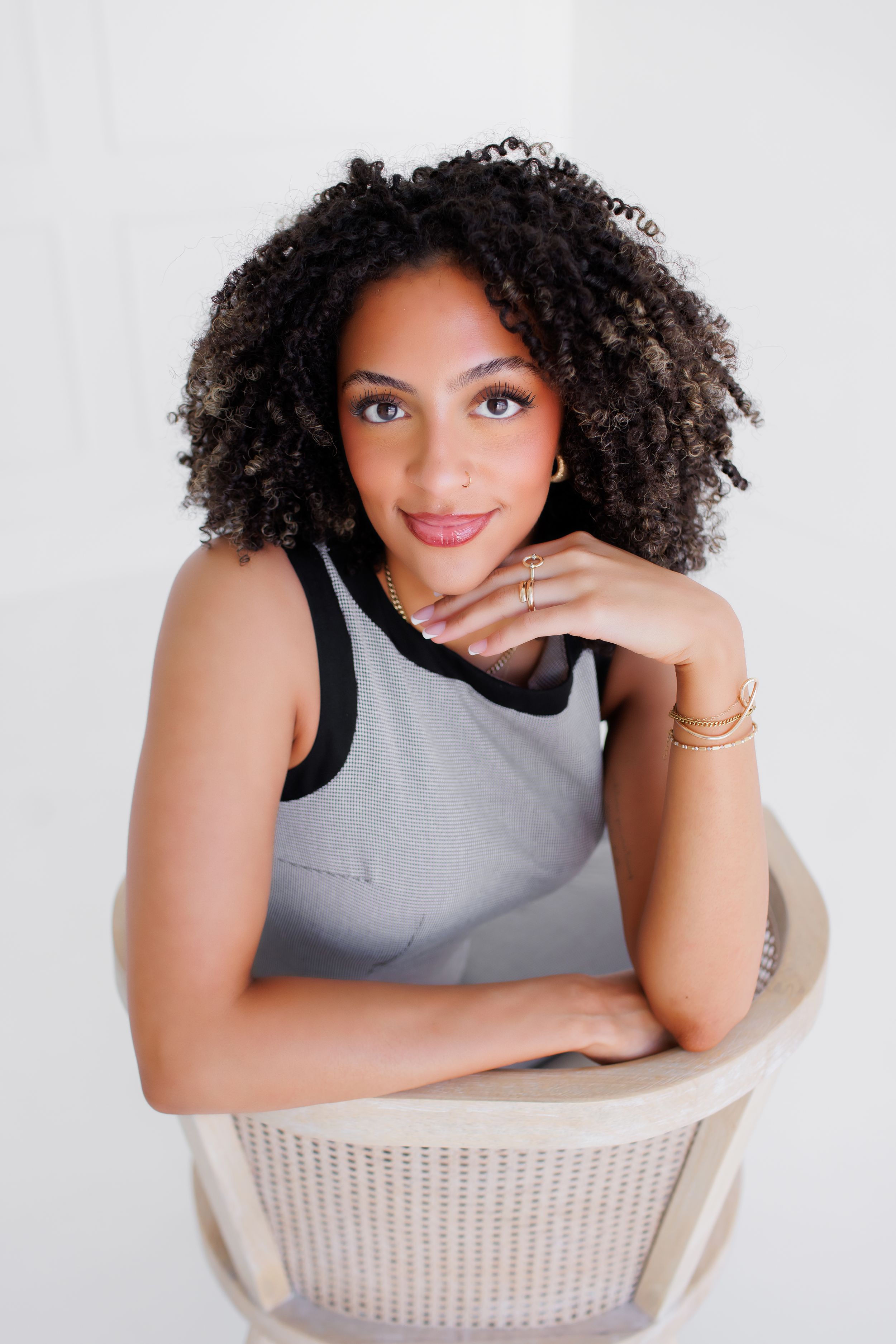 Young woman with curly hair sits relaxed in a chair, wearing a gray top with gold jewelry, smiling softly.