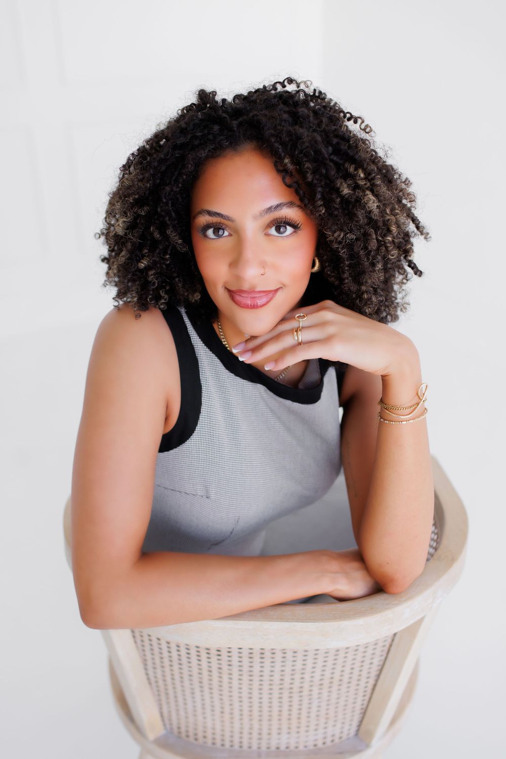 Young woman with curly hair sits relaxed in a chair, wearing a gray top with gold jewelry, smiling softly.