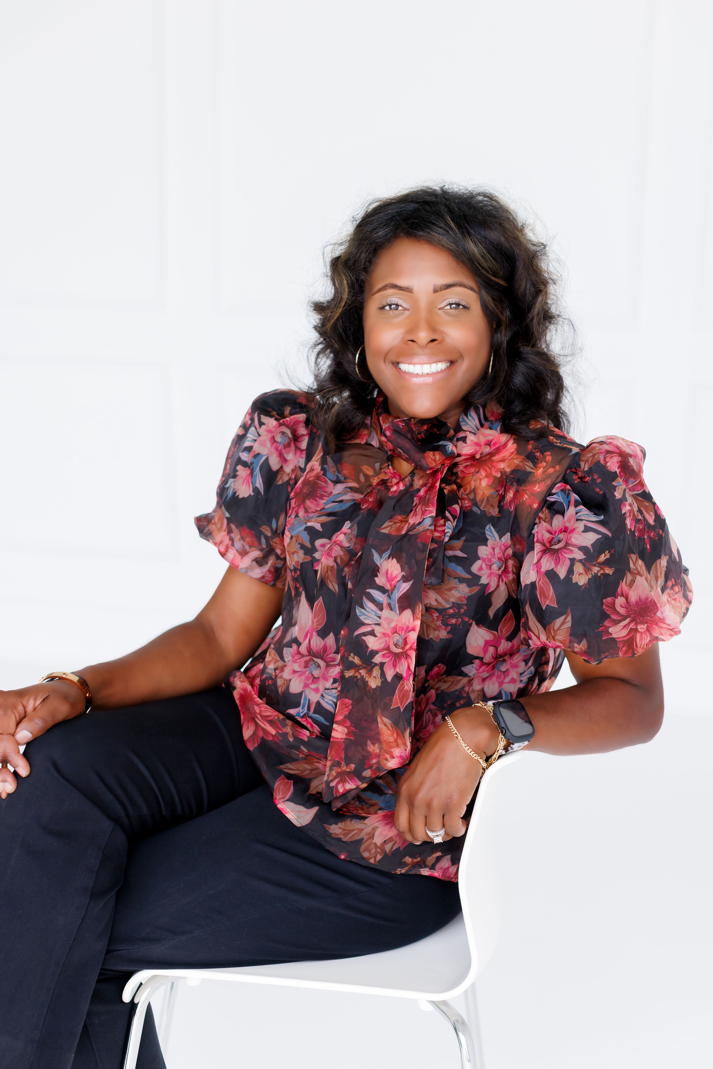 Professional photoshoot of someone wearing a pink floral ruffled blouse with black pants while posing on a white chair.