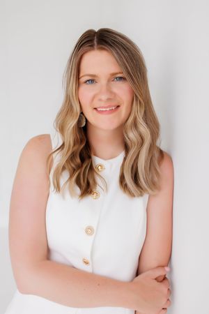 Smiling young woman with blonde wavy hair wearing a white sleeveless top with gold buttons, posing against a light background.