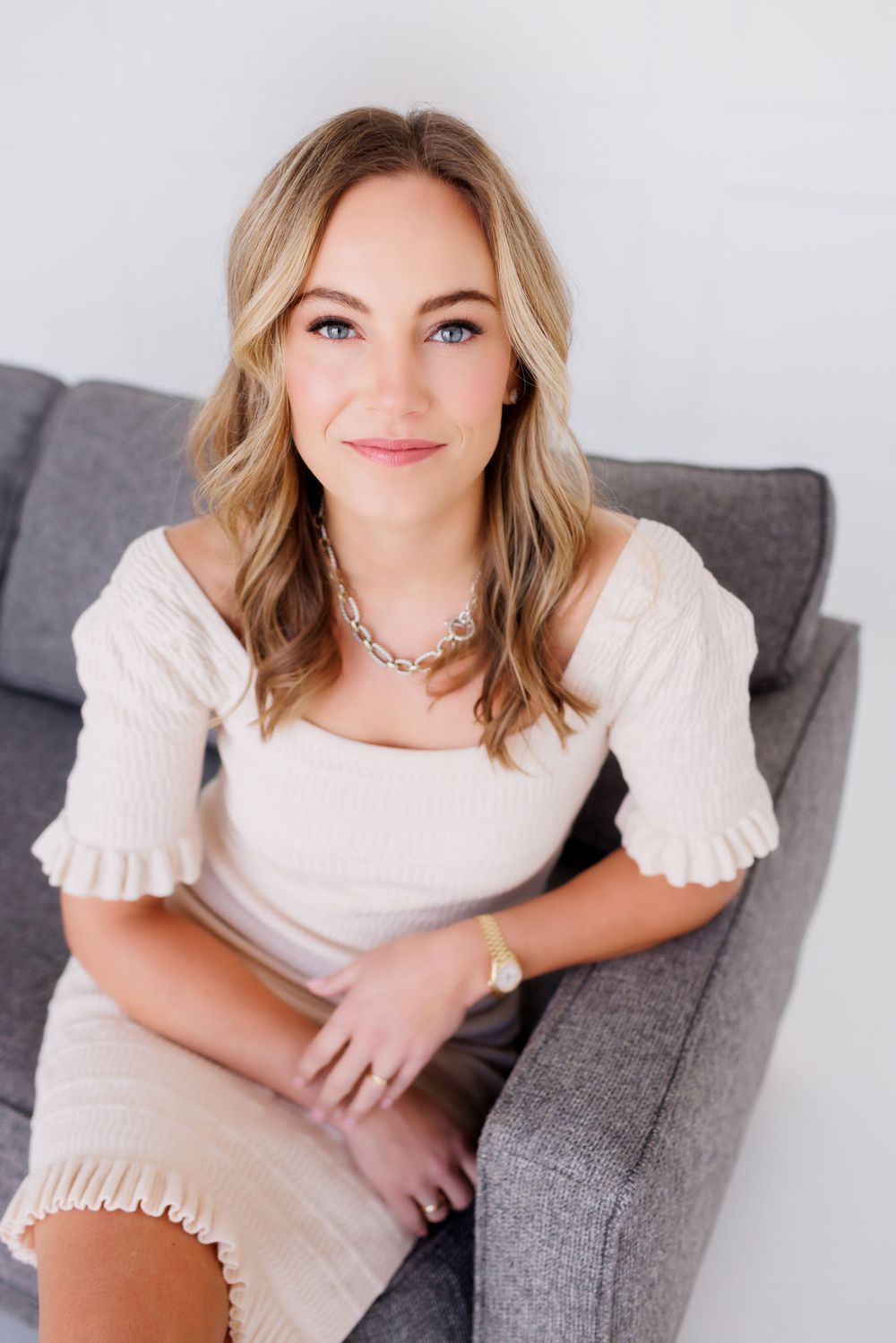 A woman in a cream-colored ruffle dress sits casually on a gray couch while wearing a necklace.