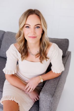 A woman in a cream-colored ruffle dress sits casually on a gray couch while wearing a necklace.