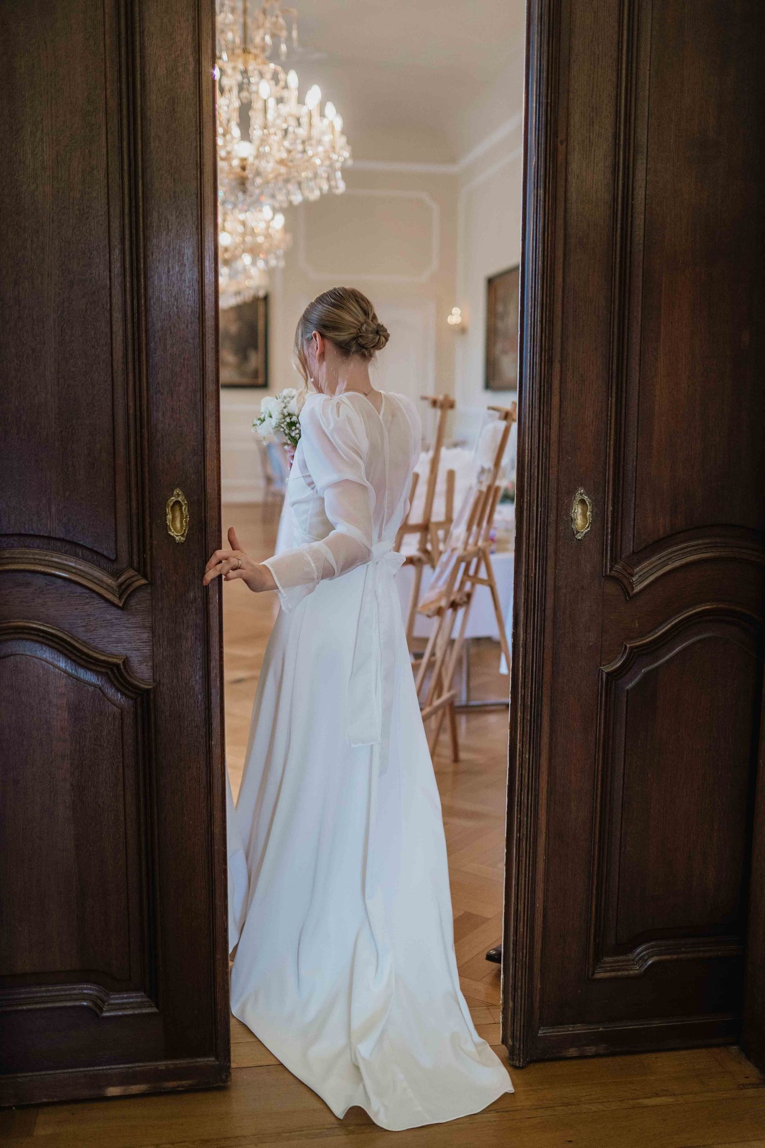 Elegant bride in long-sleeved wedding gown standing in doorway of ornate room with crystal chandelier.