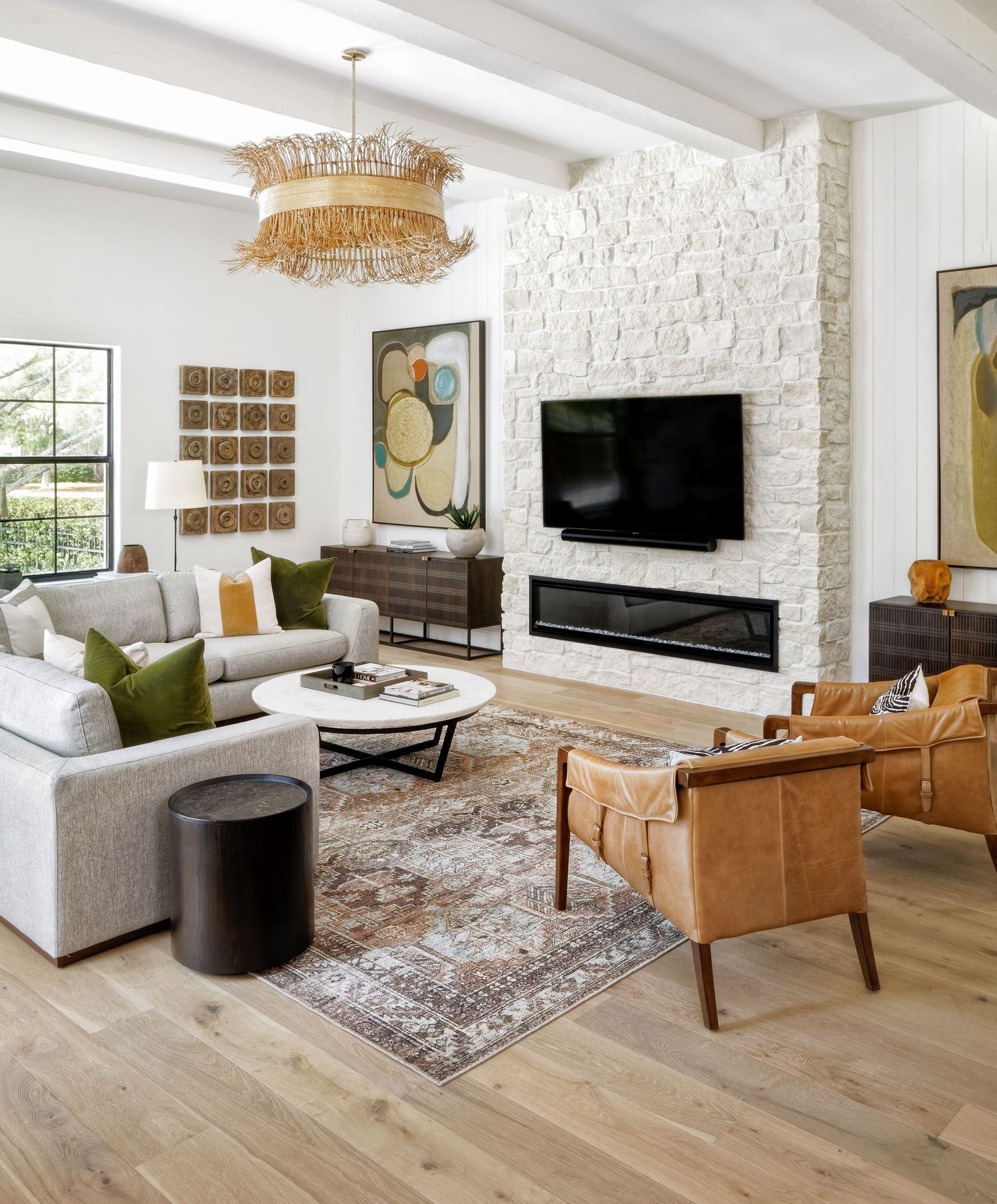 Bright farmhouse living room with white stone fireplace, leather chairs, gray sectional, and woven rattan chandelier.