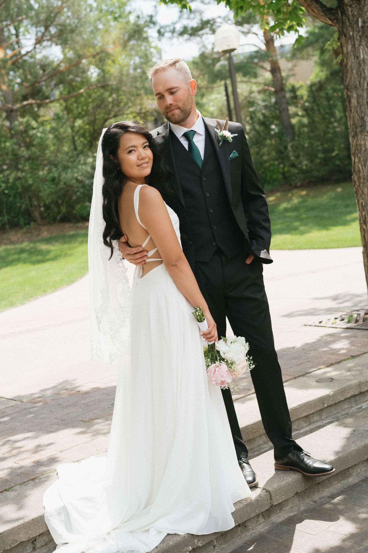 A happy couple in formal wedding attire pose together outdoors under trees on a sunny day with green foliage in background.
Saskatoon photo