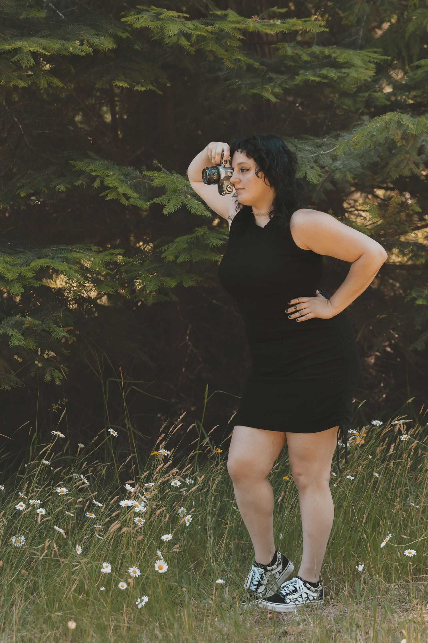 A woman standing in a field of daisys with a tree behind her. She is holding a camera. She is wearing checkered pants and a black shirt.