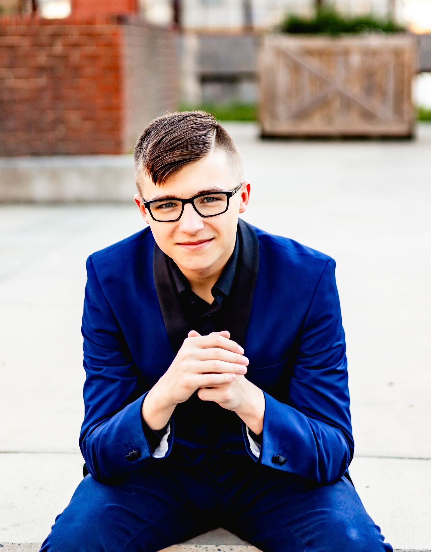 Professional headshot of a person in a navy blue blazer and black shirt sitting with clasped hands.