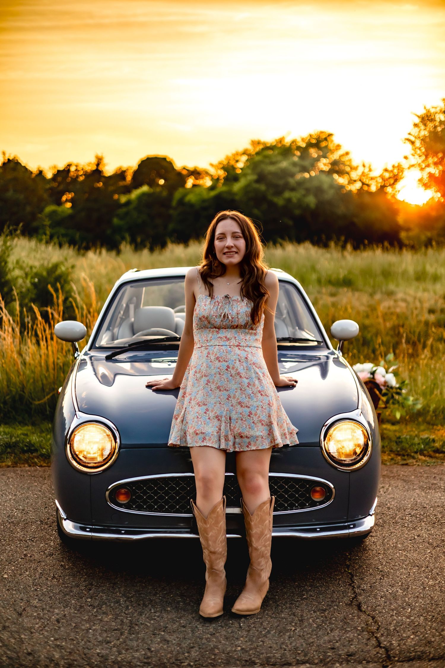 Person in floral dress and boots poses in front of classic car at sunset.