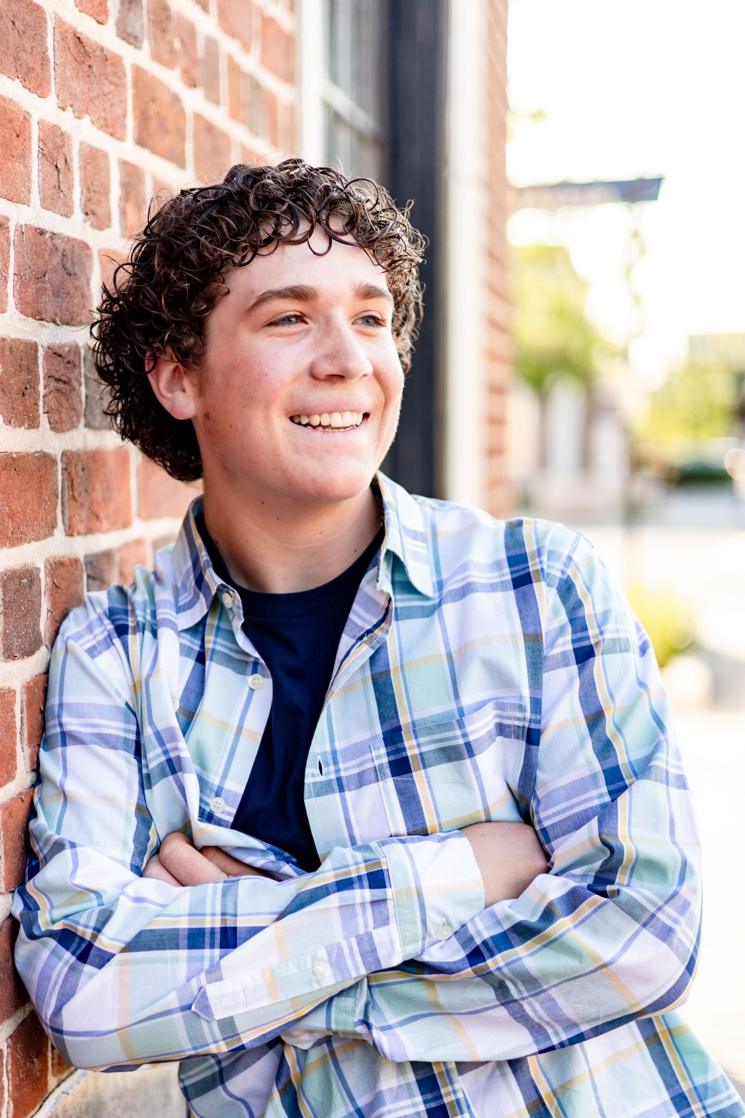 Person in plaid shirt leaning against brick wall smiling at camera.