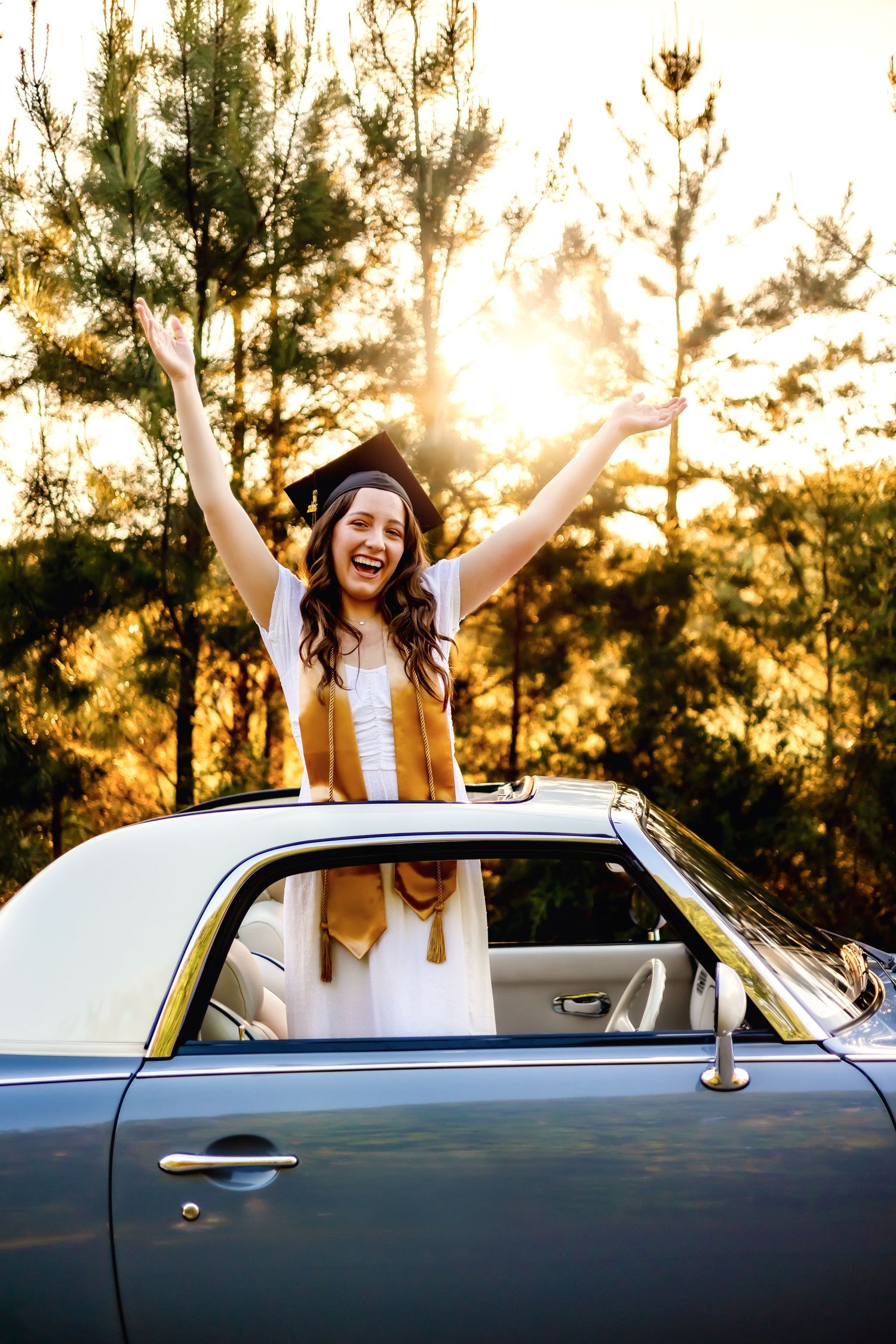 Person celebrating in vintage car with arms raised during golden hour sunset.