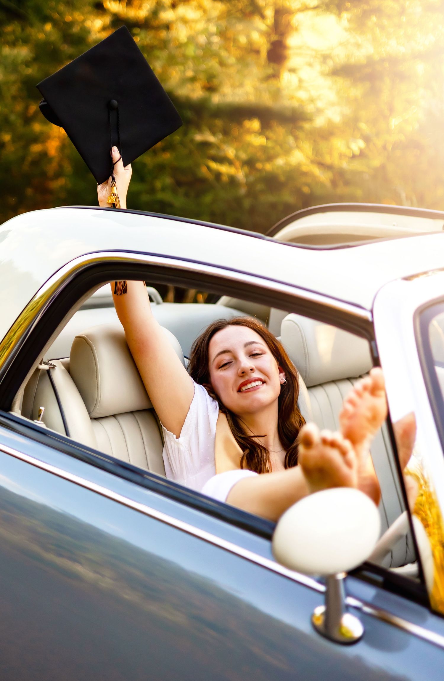 Graduate holding up cap while celebrating in car during sunset.