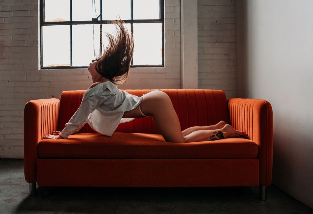 Person laying on a vibrant orange couch with windswept hair near a window in an industrial-style room.