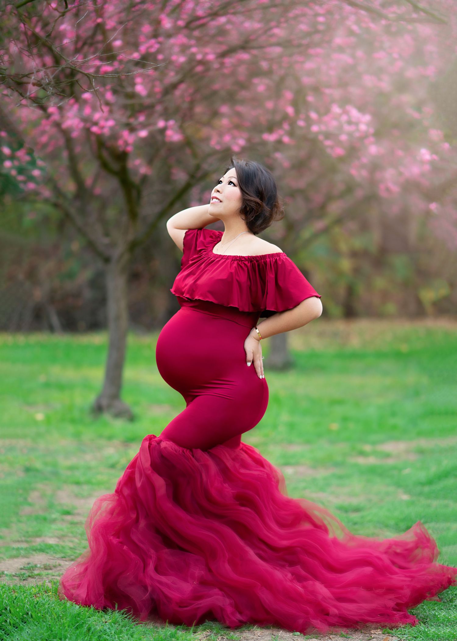 Maternity portrait in pink flowing gown against floral backdrop in moody lighting.