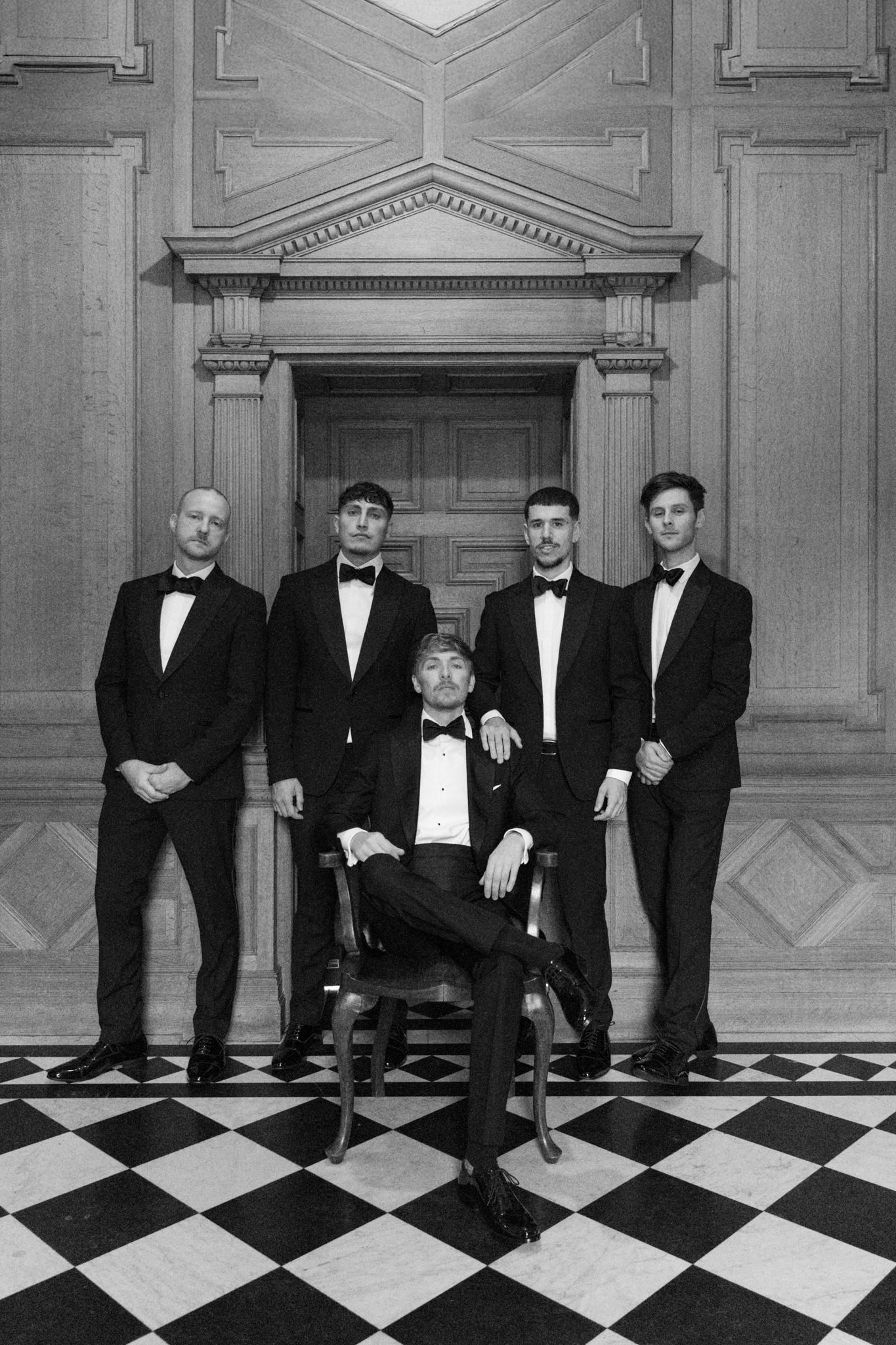 A groom and his 4 groomsmen posing in tuxedos on a chequerboard floor