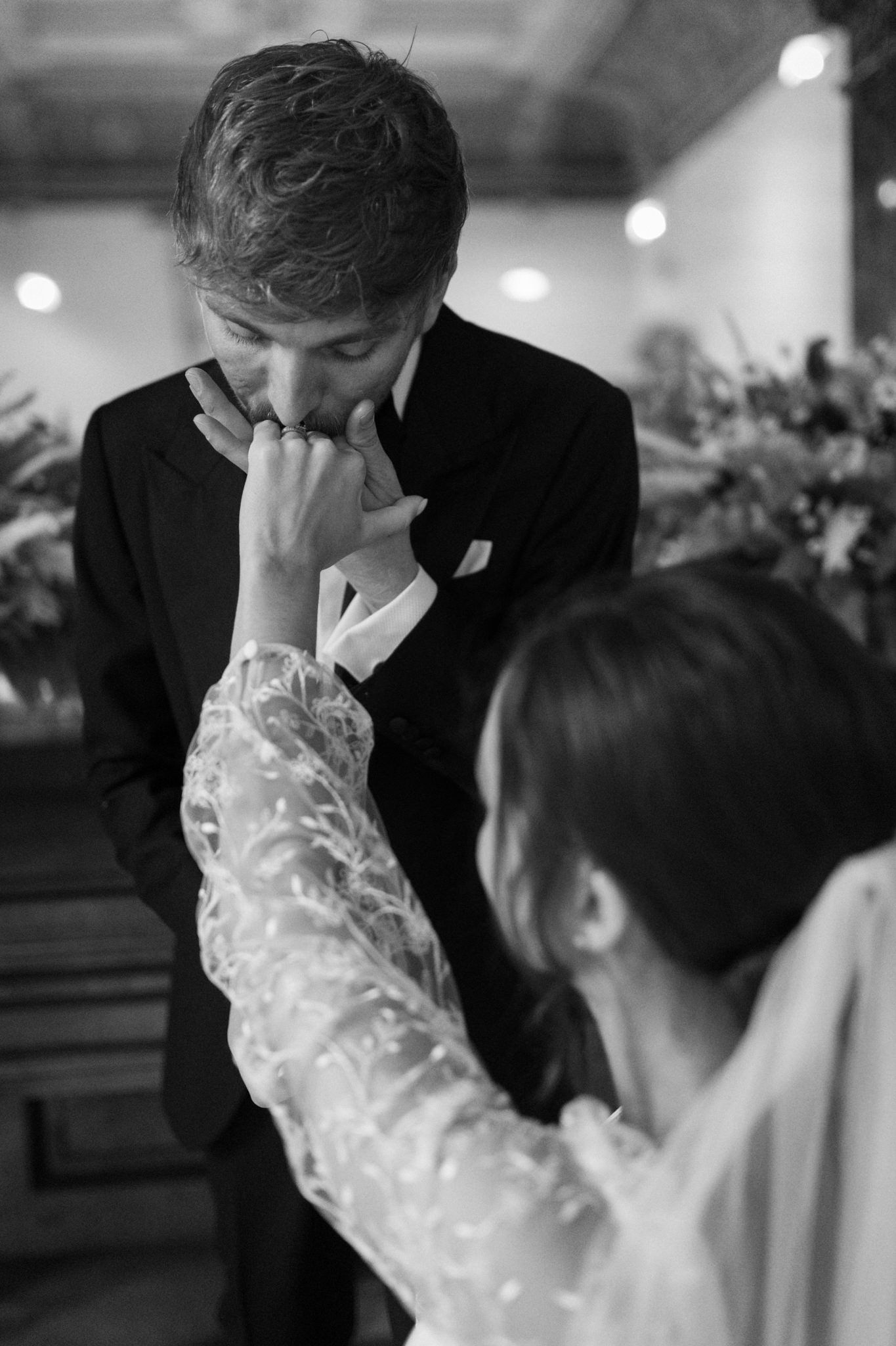 black & white image of a groom in a tuxedo kissing his brides hand as she sits in front of him