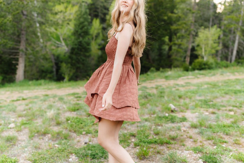 Person in dusty rose dress walking through mountain meadow with evergreen trees.