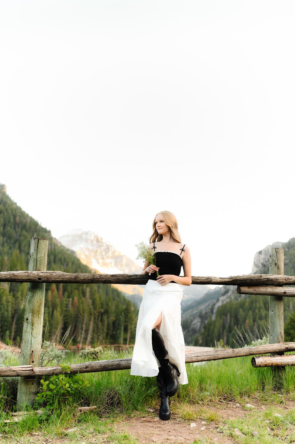 A bride in a white dress reclines on a rustic wooden fence with mountain scenery in the background.