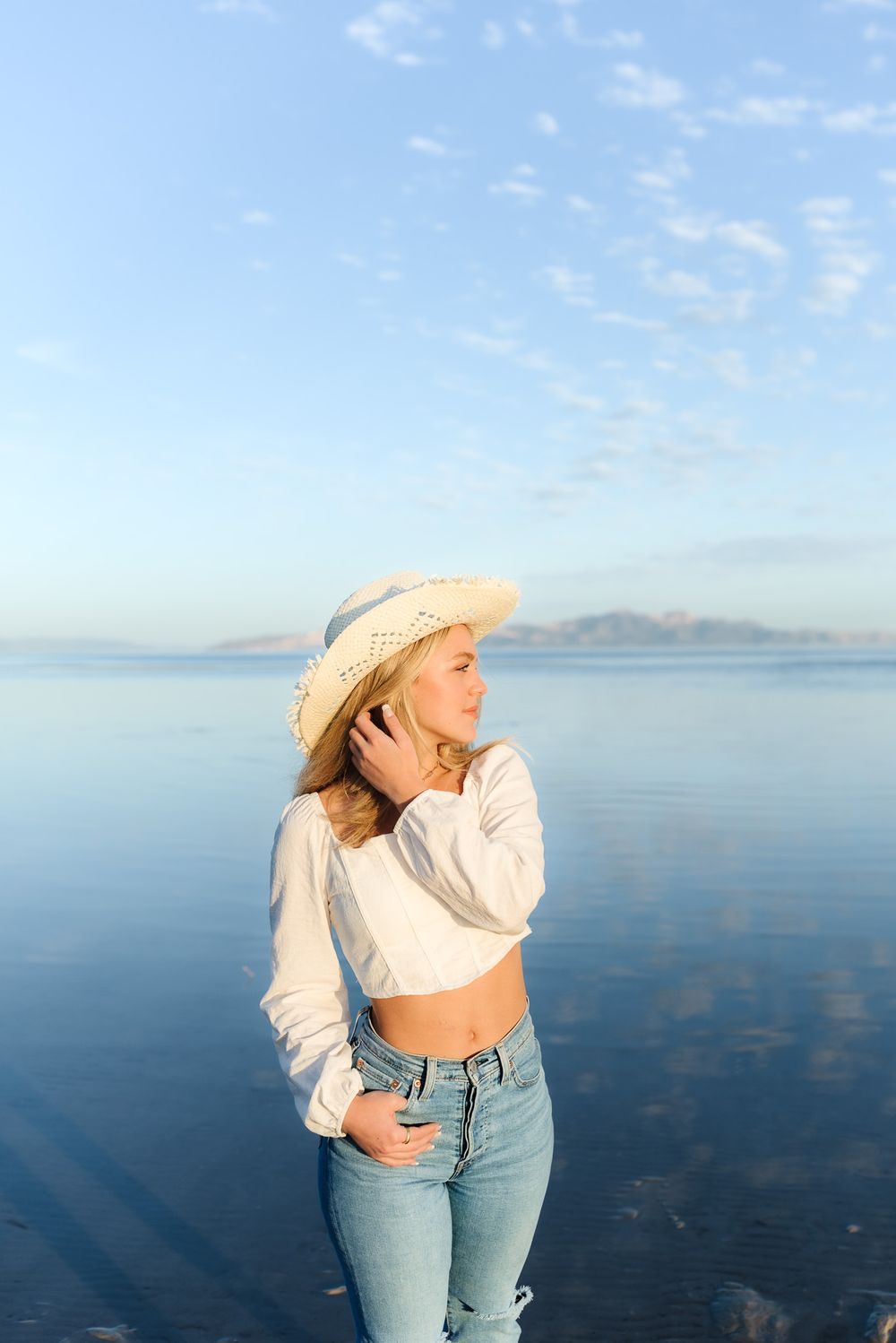 Person in white top and jeans poses against serene blue ocean backdrop with mountains in distance.