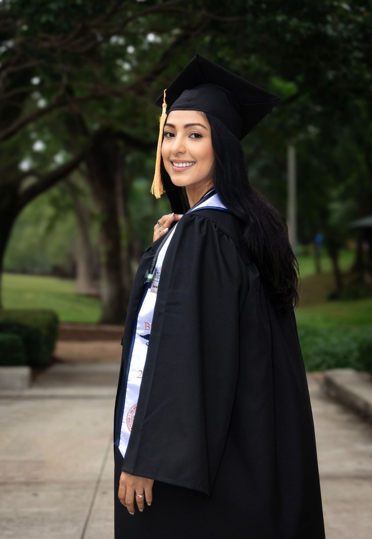 Graduate in traditional black cap and gown smiling outdoors on campus pathway.
