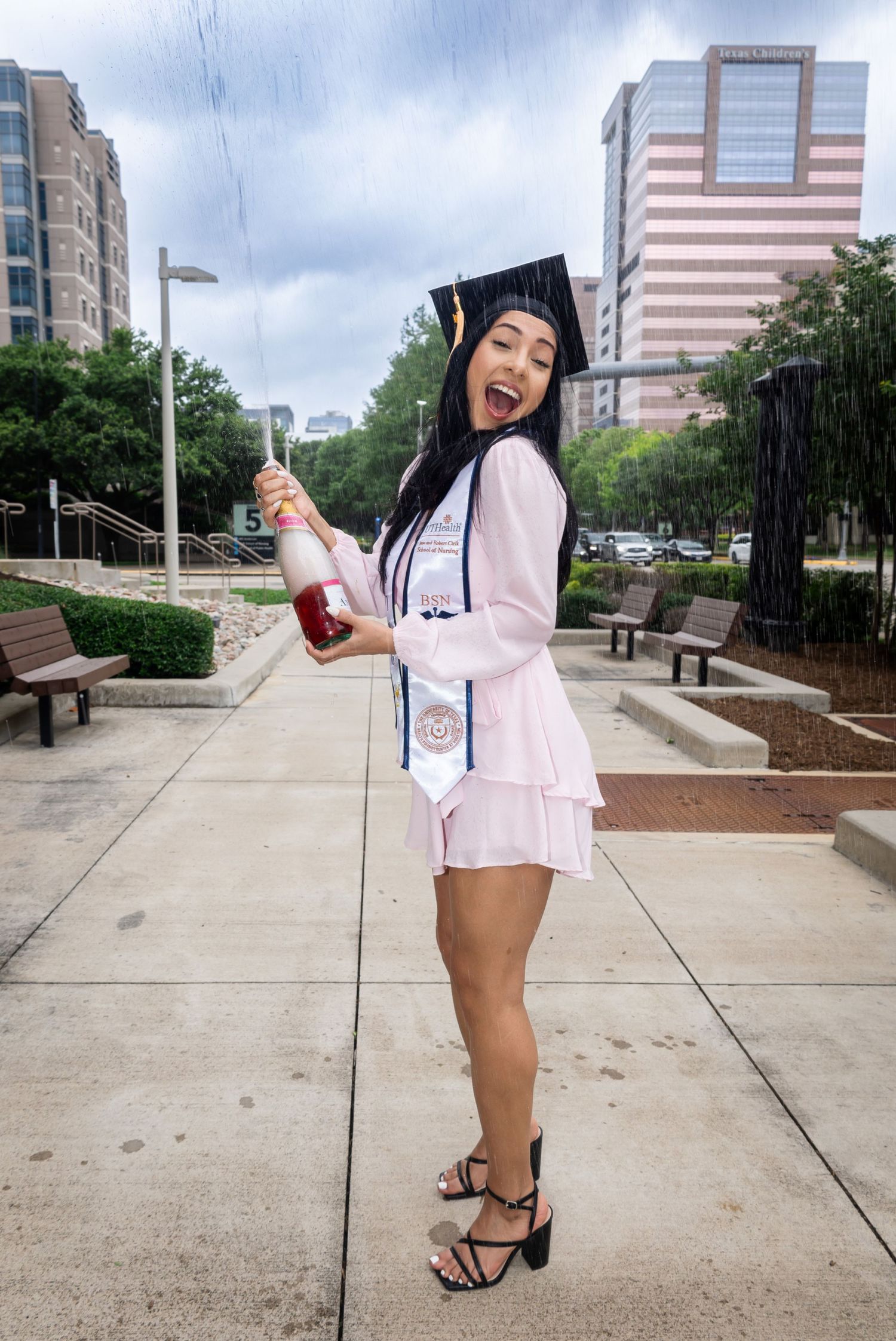 Graduate in pink dress celebrates with champagne outside urban building.