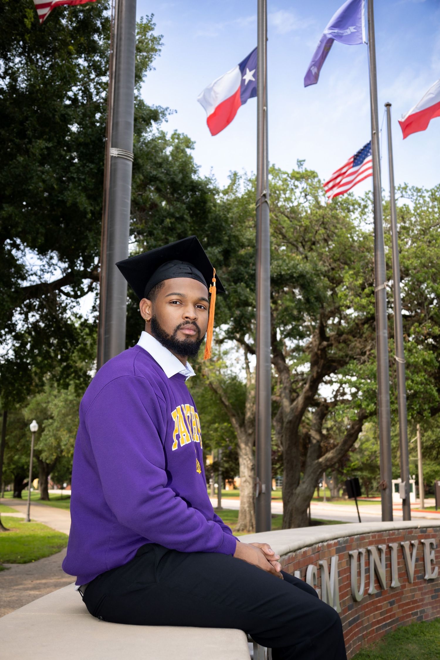 Graduate in purple sweater sits on university sign under flags.