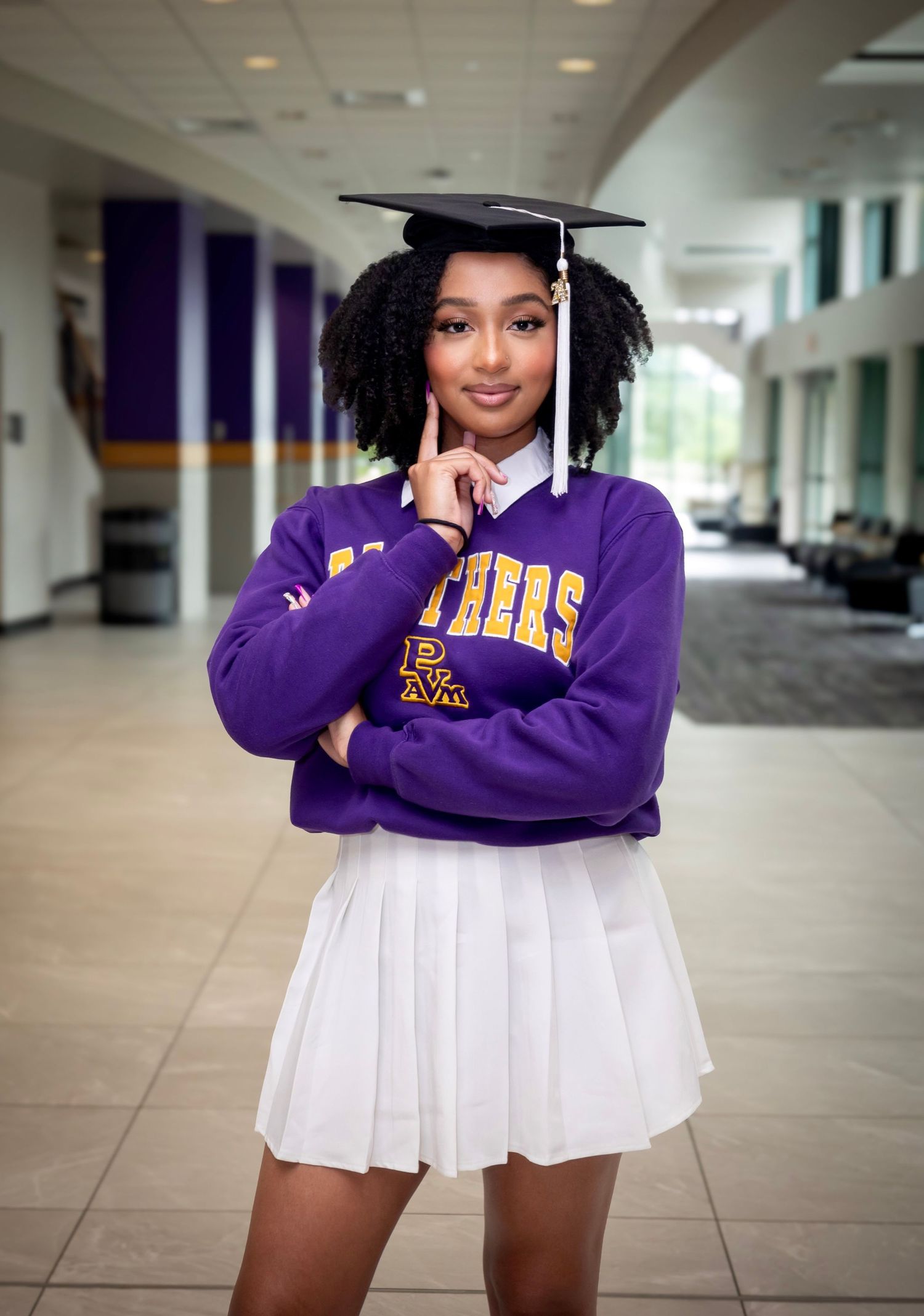 Graduate in purple university sweatshirt and white skirt posing with graduation cap.