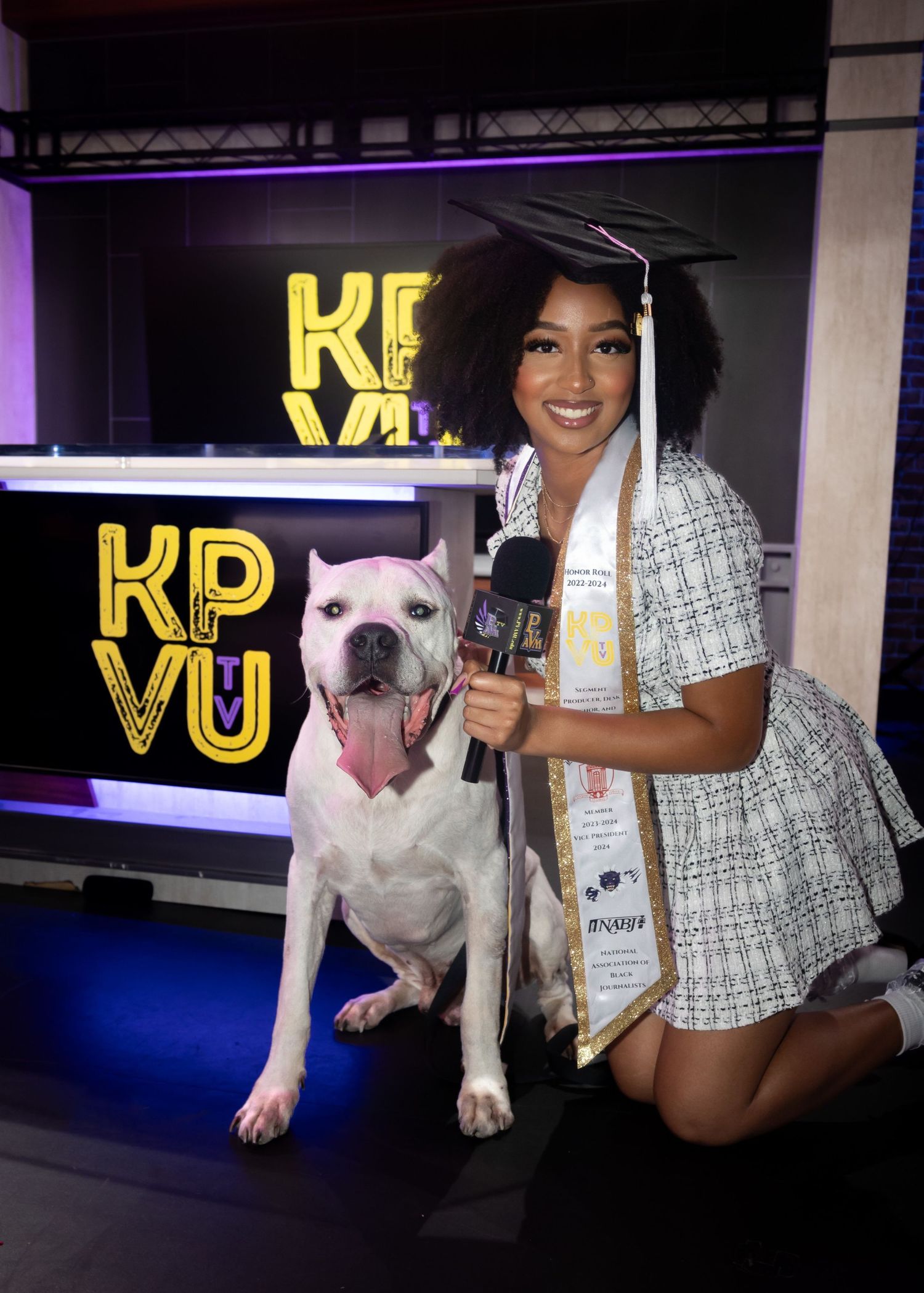 Graduate in white dress poses with white pit bull dog in front of KP VU sign.