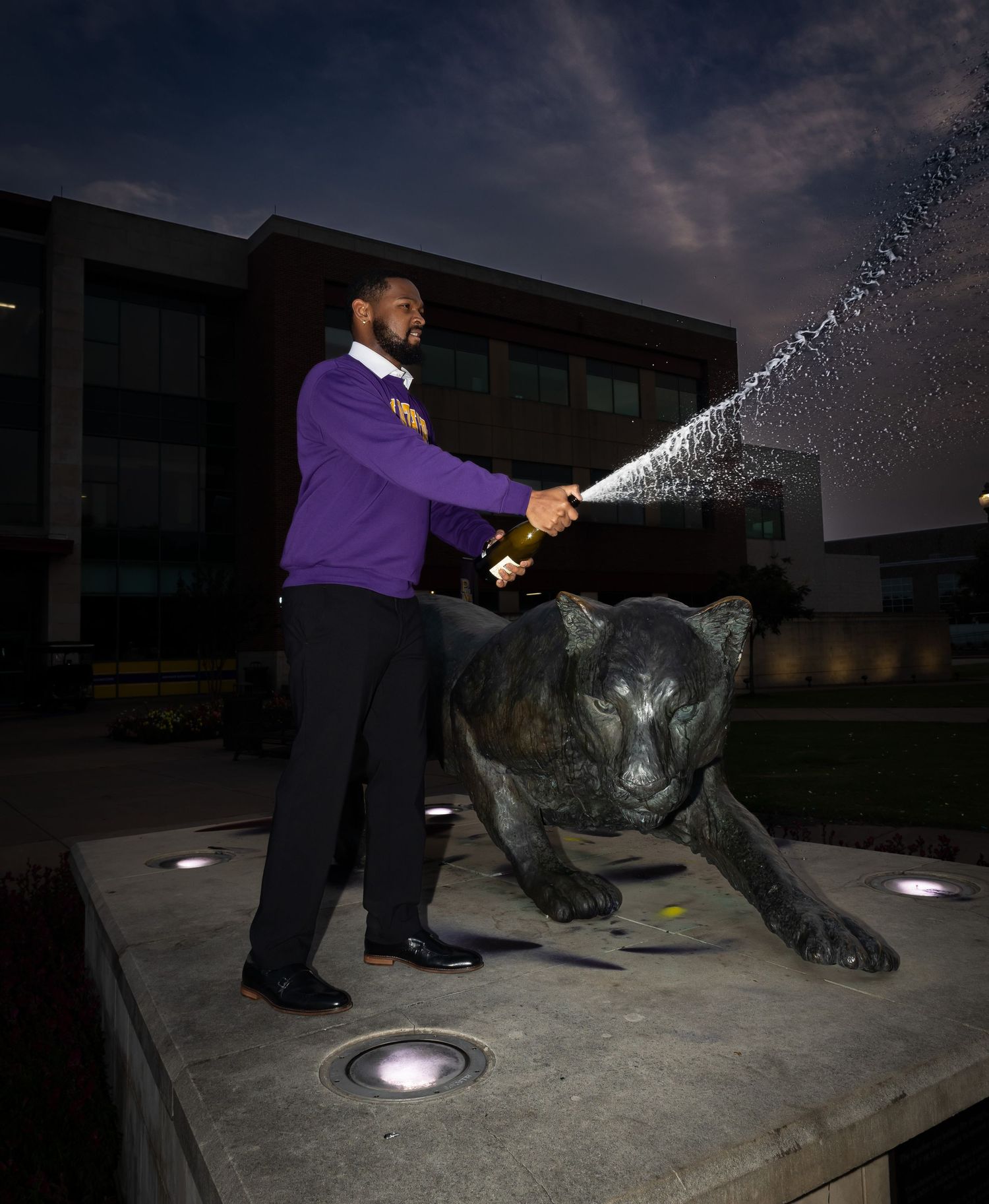 Person in purple sweater sprays champagne next to large panther statue at night.