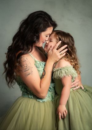 Mother and child share a tender moment wearing matching sage green tulle dresses against a muted background.