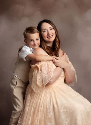 A woman in a peach gown poses with a young boy in a beige suit hugging her from behind in a studio portrait.