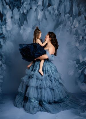A dreamy portrait in blue tones shows two figures in flowing tulle gowns against a textured floral backdrop.
