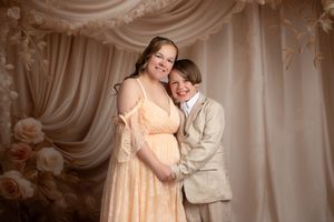 A woman in a peach dress and a boy in a tan suit pose together smiling before a floral draped backdrop.