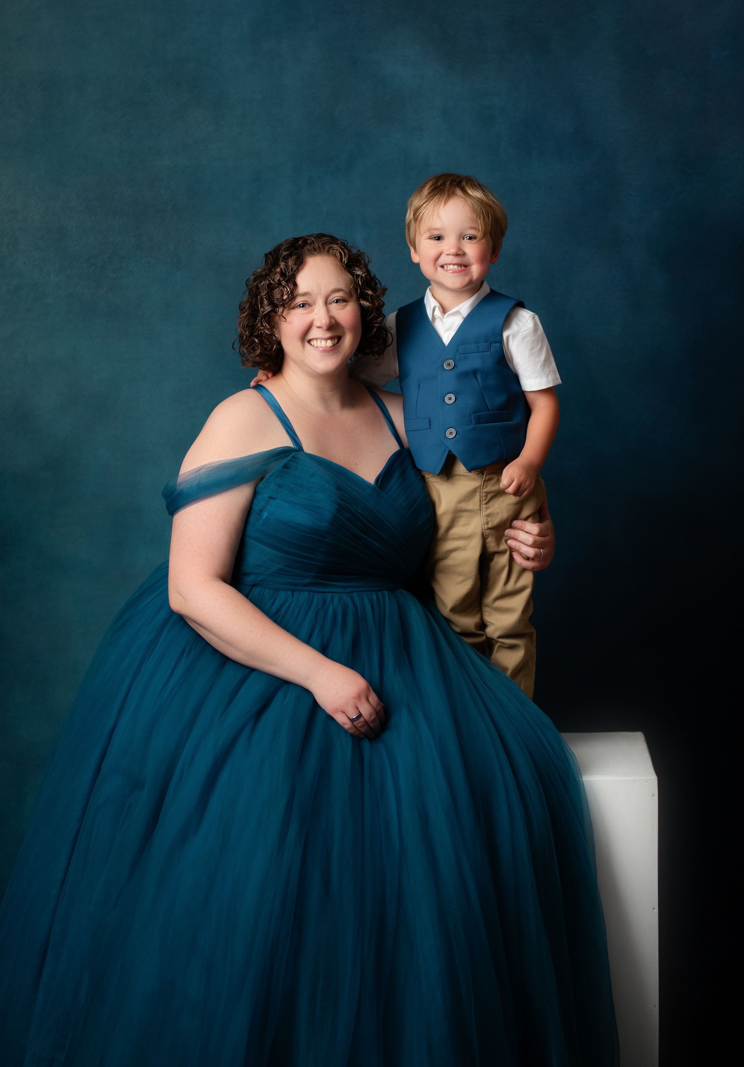 Woman in teal ball gown poses with smiling young boy in blue vest and khakis against blue backdrop.