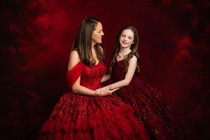 Two people in elegant red dresses share joyful moments against a dark background during a photoshoot.