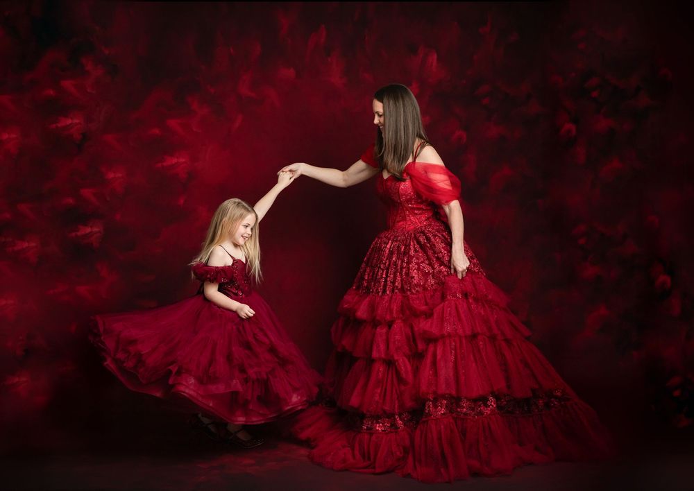 Two figures in matching red ruffled gowns pose together against a dark red background in an elegant portrait.