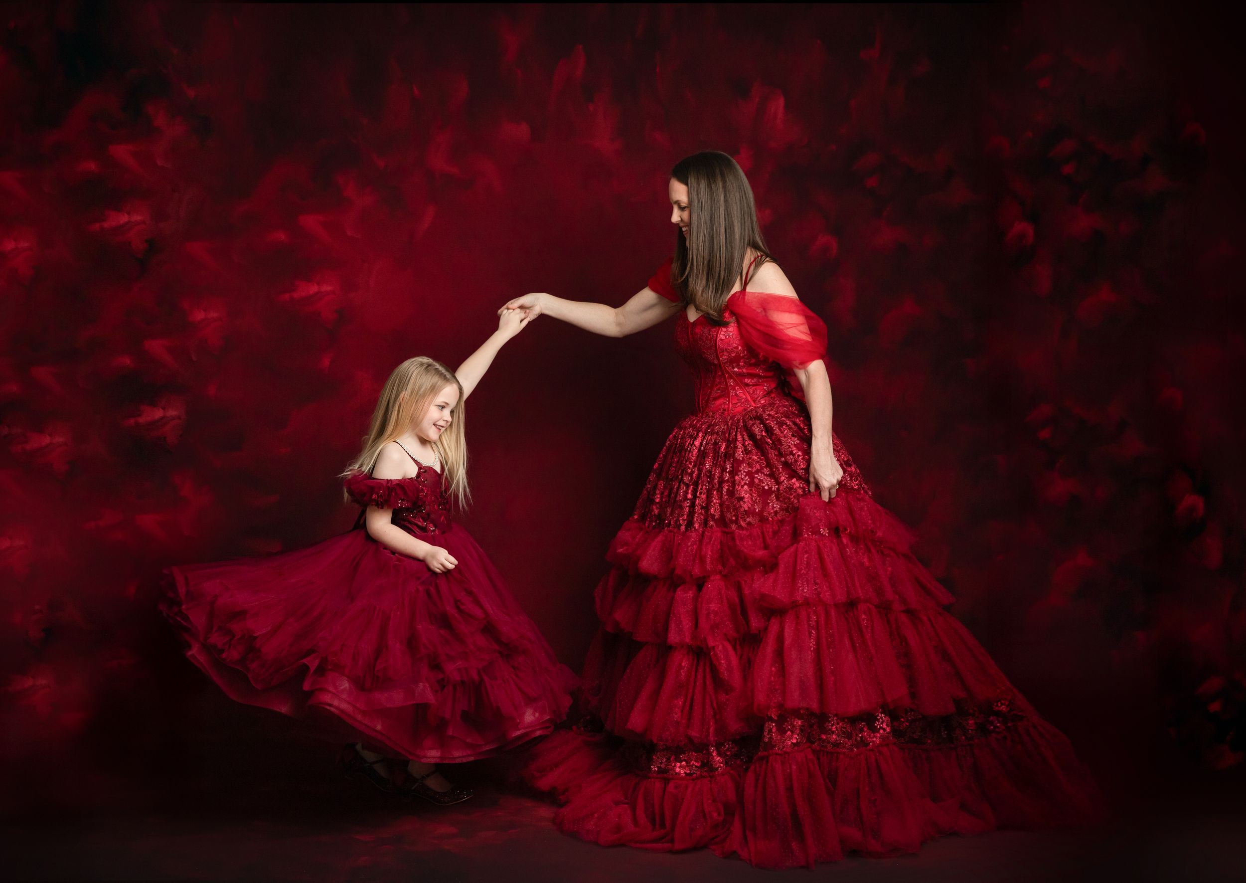 Two figures in matching red ruffled gowns pose together against a dark red background in an elegant portrait.