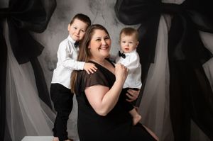Family portrait featuring a woman in a black dress with two young boys against a gray and white draped background.