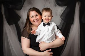 A person in brown holds a young child wearing a white dress shirt and bow tie during a formal photo session.