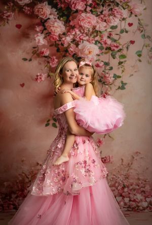 Two figures in pink floral dresses pose together against a romantic rose-covered backdrop in a dreamy portrait session.