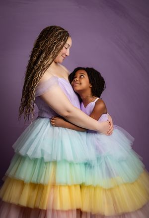 Two people wearing matching pastel rainbow tulle dresses embrace in a sweet moment against a purple backdrop.