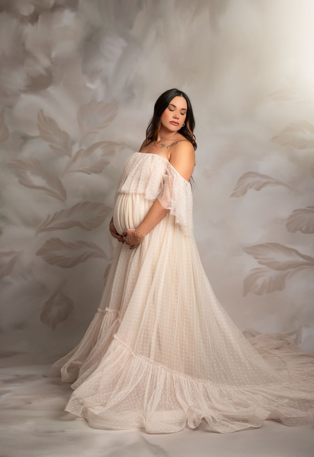 Expecting mother poses in flowing off-shoulder cream gown with delicate layers against floral backdrop.
