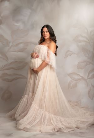 Ethereal maternity portrait in flowing white dress against soft gray floral background.