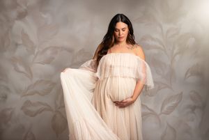 A pregnant figure poses in an ethereal off-shoulder cream gown against a textured backdrop with leaf patterns.
