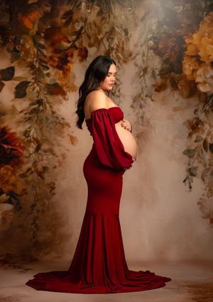 A pregnant woman in a flowing burgundy off-shoulder gown poses elegantly against an autumn-themed backdrop with floral elements.