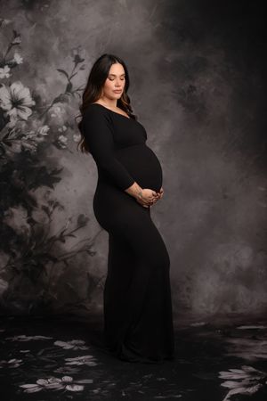 A moody maternity portrait shows an expectant mother in a black dress against a dark floral backdrop.