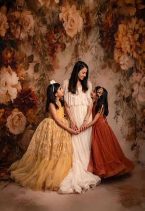 Three women in elegant vintage-inspired dresses sit together against a moody floral backdrop in warm earth tones.