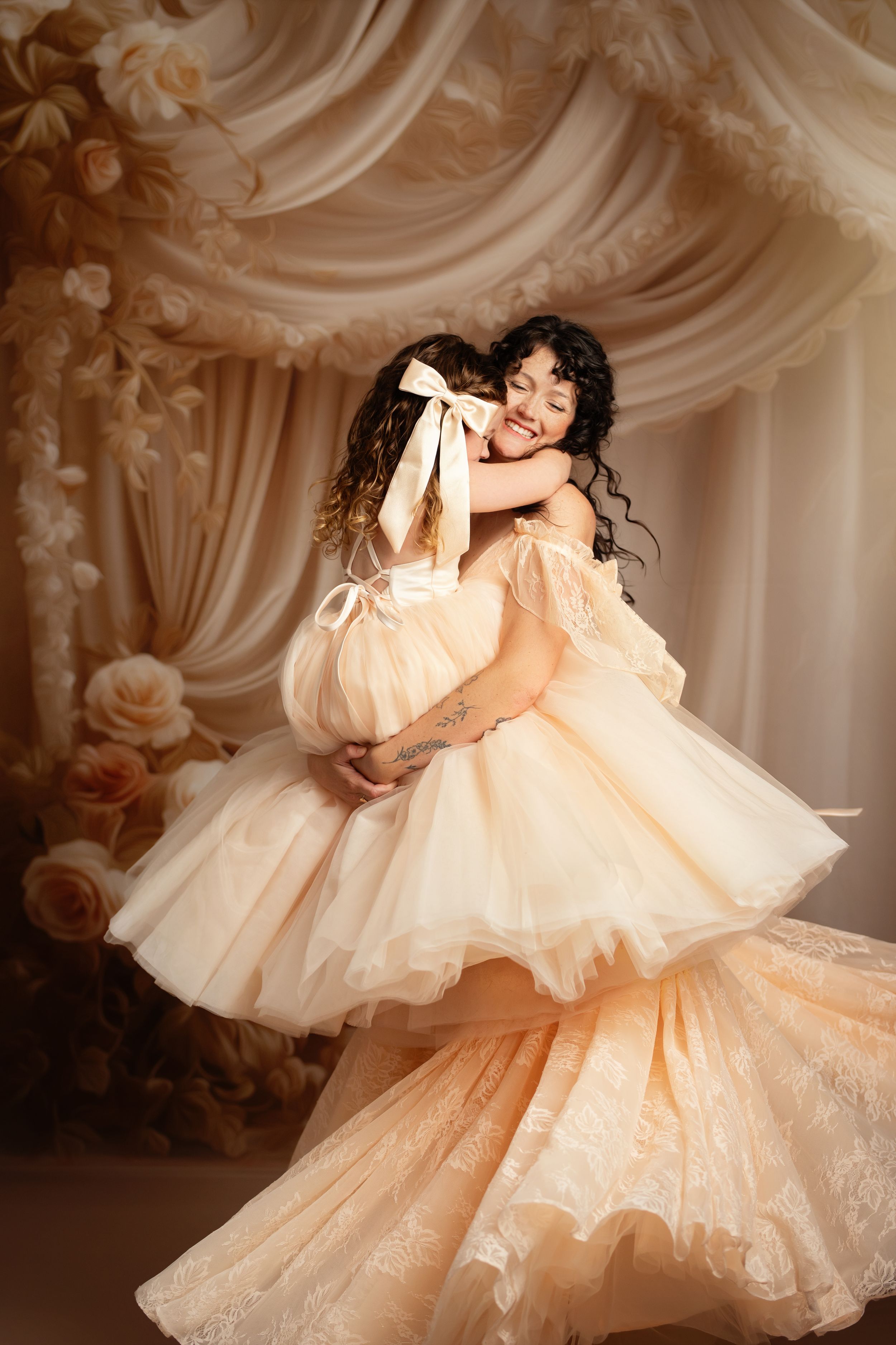 Mother and daughter in matching peach tulle gowns share a joyful embrace in an elegant floral studio setting.