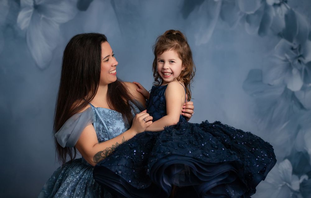 A mother and daughter share joyful moments in matching blue sparkly dresses against a moody gray backdrop.